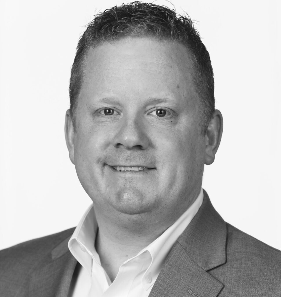 Professional headshot of Sean Corcoran, a man with short curly hair and a trimmed beard, smiling slightly at the camera, wearing a dress shirt and gray suit jacket against a plain white studio background, in black and white